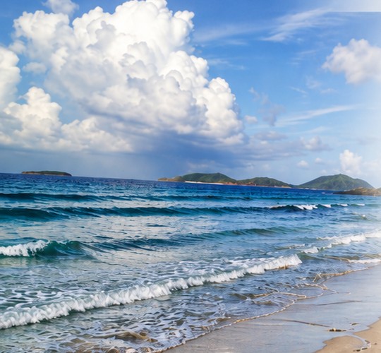 Turquoise waves meeting a sandy shore, with green islands under a bright sky.